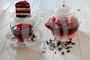 Hibiscus tea in cup and teapot, tea leaves, piece of red velvet cake on white wooden background.