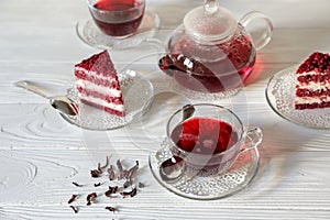 Hibiscus tea in cups and teapot, tea leaves, for dessert red velvet cake pieces on white wooden background.