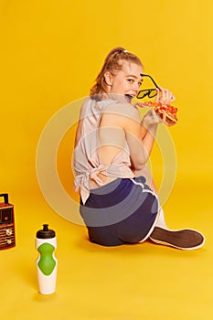 Hiding food, secrets. Young beautiful girl in sunglasses and sports wear eating pizza, posing over yellow studio