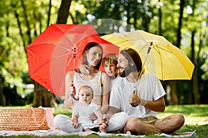 Hiding from the sun under big red and yellow umbrellas mother, father and their children are sitting on the blanket and