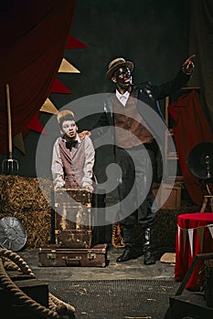 High African man with dirty face and clothes standing with little servant boy over dark retro circus backstage