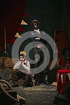 High African man with dirty face standing with little servant boy over dark retro circus backstage background. 20th