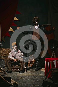 High African man with dirty face standing with little servant boy over dark retro circus backstage background. 20th