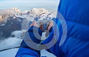 High altitude female mountaineer dressed blue warm dawn jacket holding metal mug of hot tea and drinking warm drink with