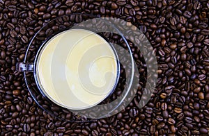 High angle close up view on isolated transparent coffee cup on glass saucer with cafe crema on countless roasted beans background