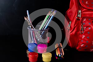 High angle closeup shot of a pen stand placed with scholl bag, crayon, pencil and oil colors against black background