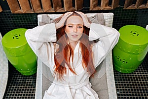 High-angle portrait of cheerful young woman in white bathrobe relaxing with closed eyes lying on deck chair after beauty