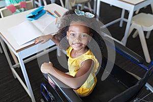 High angle portrait of smiling african american elementary girl sitting on wheelchair in classroom