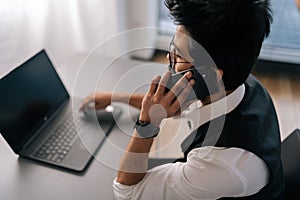 High-angle rear view of Indian bearded business man in glasses working at laptop sitting at desk and talking on