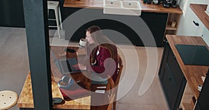 High angle shot of beautiful happy young Caucasian freelancer woman using laptop to work at home table sipping coffee.