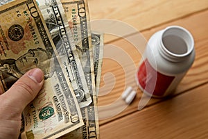 High angle shot of a person holding some dollar bills over a bottle of pills on a wooden surface