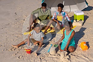 High angle view of african american parents sitting under umbrella while children playing with sand