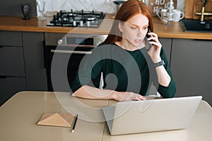 High-angle view of angry female freelancer talking on mobile phone and using laptop sitting at table in kitchen with