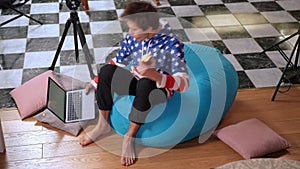 High angle view bag chair indoors with Caucasian confident boy entering surfing Internet on laptop eating apple. Teenage