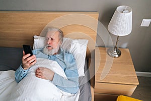 High-angle view of bearded mature adult male using smartphone lying on bed in bedroom at home.