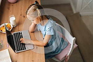 High-angle view of bored elementary child girl using typing laptop keyboard sitting at home table with snack by window.