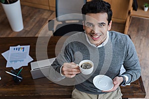 High angle view of businessman with saucer and cup of coffee smiling and looking at camera near table