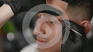 High angle view closeup young Middle Eastern man doing eyebrows shaping in beauty parlor. Caucasian unrecognizable woman