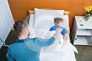 High angle view of father touching sick little son lying in hospital bed with teddy bear