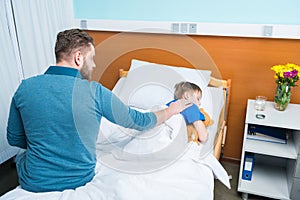 High angle view of father touching sick little son lying in hospital bed with teddy bear