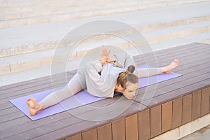 High-angle view of flexible young woman sits on twine on yoga mat performing forward, stretching in city park.