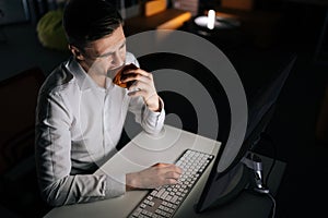 High-angle view of freelancer male eating burger working on desktop computer sitting on desk at office workplace late