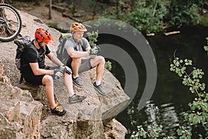 high angle view of friends cyclists with backpacks resting with sport bottle of water on rocky cliff near river