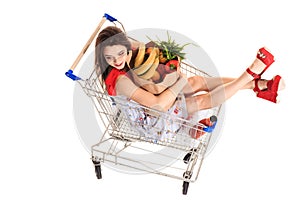 High angle view of girl smiling at camera while holding grocery bags and sitting in shopping trolley isolated on white