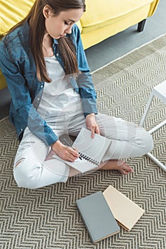 high angle view of girl writing in notebook while sitting on carpet and studying