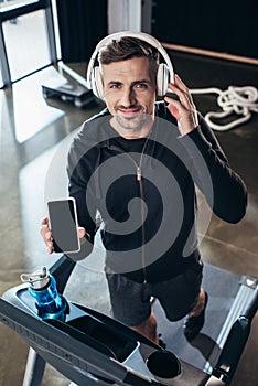 high angle view of handsome sportsman in hoodie showing smartphone with blank screen on treadmill