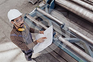 High angle view of heavy industry worker with safety headphones and hard hat in industrial factory holding blueprints