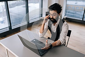 High-angle view of Indian bearded businessman in glasses working at laptop computer sitting at desk and talking on