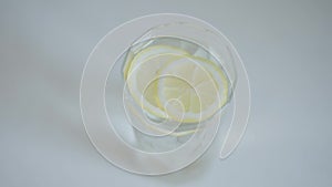 High angle view lemon slices dropping in glass of water. Close-up healthful vitamin drink on white table indoors