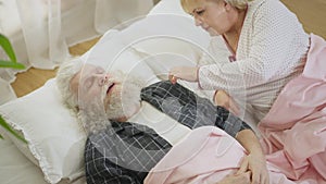 High angle view of old Caucasian man snoring sleeping in bed waking up woman. Dissatisfied sad wife touching shoulder of
