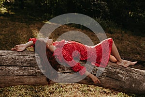 High angle view portrait a young and beautiful woman in a red summer dress with white flecks resting on a fallen dry tree trunk in