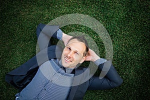 Happy Young businessman in a suit lying on the green grass and relaxing in park.