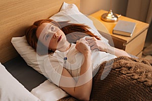 High-angle view of sick young woman with flu lying under blanket on bed holding thermometer in armpit. Ill female with