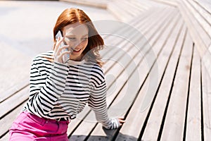 High-angle view of smiling redhead female hipster talking by mobile phone outdoors sitting on bench in city park. Happy