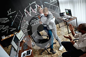 High-angle view of three traders celebrating their success while working in the office. Blackboard full of charts and