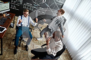 High-angle view of three traders celebrating their success while working in the office. Blackboard full of charts and
