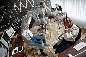 High-angle view of three traders celebrating their success while working in the office. Blackboard full of charts and