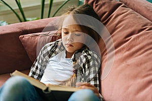 High-angle view of tired adorable child kid girl reading paper book lying on soft couch at home looking at camera.