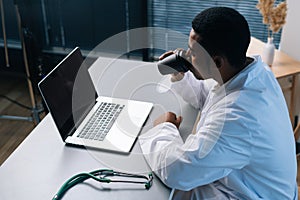 High-angle view of tired black male doctor wearing white coat uniform using working on laptop computer sitting at desk