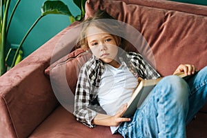 High-angle view of unhappy adorable child kid girl reading paper book lying on soft couch at home looking at camera.