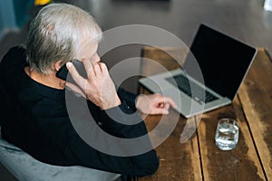 High-angle view of unrecognizable gray-haired senior adult man talking on mobile phone sitting at desk with laptop