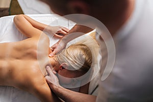 High-angle view of unrecognizable male masseur doing back and neck massage to naked female client in spa salon lying on