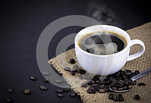 High angle view of white ceramic cup of black hot Americano coffee with steam on a sackcloth with a pile of roasted coffee beans