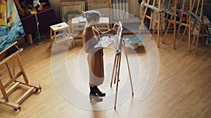 High angle view of young woman painter working in studio standing in front of easel and painting holding brush and