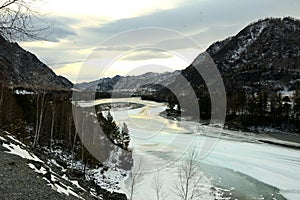 The high bank of a beautiful river flowing through a picturesque mountain valley with snow-covered slopes on a cloudy December