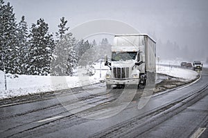 High cab long hauler white big rig semi truck transporting cargo in dry van semi trailer slowly drive on the wet winter highway